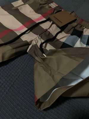 Burberry shorts  - Säljer ett par klassiska badshorts från Burberry med det ikoniska rutmönstret i beige, brunt, svart, vitt och rött. Shortsen har resårmidja med dragsko och innerbyxa i mesh. Perfekta för sommaren och strandhäng.