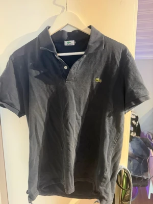 Svart pikétröja från Lacoste - Size XL men passar S. Klassisk svart pikétröja från Lacoste med korta ärmar och den ikoniska gröna krokodilloggan på bröstet. Tröjan har krage och knappar vid halsen. Perfekt för en clean och stilren look.