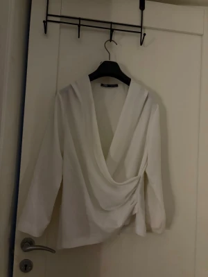 Vit omlottblus med drapering - Stilren vit omlottblus med djup v-ringning och snygg drapering framtill. Blusen har långa ärmar och rak passform. Perfekt att matcha med jeans eller kjol för en trendig look. Materialet ser ut att vara ett mjukt tyg med lätt struktur.