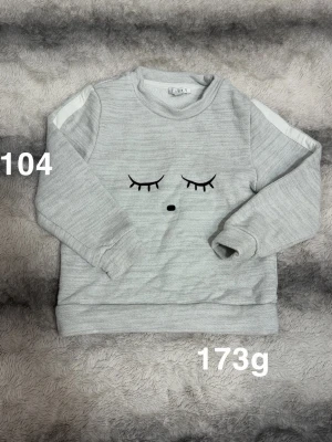 Ljusgrå sweatshirt med ögonmotiv - Ljusgrå sweatshirt med rund hals och långa ärmar. Framtill finns ett broderat motiv av stängda ögon och en liten näsa. Tröjan har vita detaljer på axlarna och ribbade muddar vid ärmslut och nederkant. Perfekt för en avslappnad stil.🌸 Fint använt skick. Storlek 104
