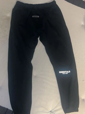 Svarta Essentials mjukisbyxor - Säljer ett par svarta mjukisbyxor från Essentials med tryck på benet. Byxorna har resår i midjan och vid bensluten samt sidofickor. Perfekta för chill dagar och streetwear-stil.
