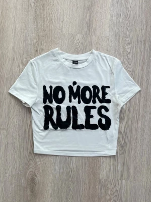 Vit croppad t-shirt med tryck - Säljer en vit croppad t-shirt med svart texten 'NO MORE RULES' framtill. T-shirten har korta ärmar och en avslappnad passform. Perfekt för dig som vill sticka ut med ett statement-plagg.