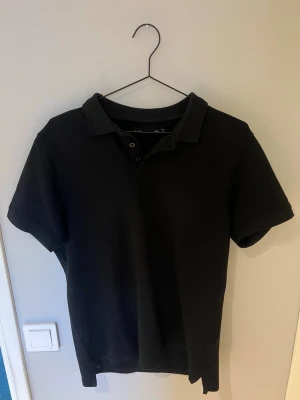 Svart piké t-shirt med krage - Klassisk svart piké t-shirt med korta ärmar och krage. Tröjan har knappar vid halsen och är tillverkad i ett mjukt bomullsmaterial. Enkel och stilren design som passar till det mesta.