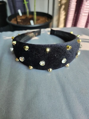 Diadem med nitar och stenar - Svart sammetsdiadem med nitar och stenar Snyggt svart diadem i sammet med guldfärgade nitar och glittrande stenar. Bred modell som ger ett statement till din look. Perfekt accessoar för dig som vill sticka ut och addera lite edge till outfiten.  Kan även hämtas I Partille  E3