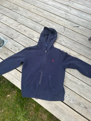 Mörkblå hoodie med dragkedja - Mörkblå hoodie med dragkedja framtill och två stora fickor. Klassisk huva och en liten röd logga broderad på bröstet. Tillverkad i mjukt bomullsmaterial, perfekt för chill dagar. Enkel och stilren design som passar till det mesta.