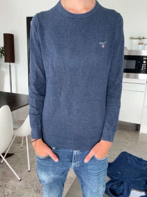 Mörkblå stickad tröja från Gant - Säljer en stilren mörkblå stickad tröja från Gant med rund halsringning och diskret logga broderad på bröstet. Tröjan har långa ärmar och ribbade muddar vid ärmslut och nederkant. Perfekt till jeans för en clean look. Tröjan är M men passar mer S