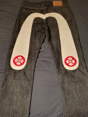 Evisu jeans med vita detaljer - Säljer ett par mörkblå jeans med stora vita paneler och röda blommönster på baksidan av benen. Jeansen har klassisk femficksdesign och cool läderpatch i midjan. Perfekta för dig som vill sticka ut med en riktigt speciell look.