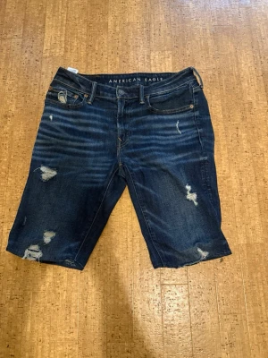 Blå jeansshorts från American Eagle - Säljer ett par blå jeansshorts från American Eagle med slitna detaljer och klassisk femficksdesign. Shortsen har en midwaist passform och är tillverkade i stretchigt bomullsmaterial (Airflex+). Perfekta för en avslappnad stil. Billigare vid snabba köp