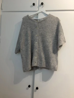 Grå kofta från Gina Tricot - Mysig grå kofta från Gina Tricot i ullblandning. Koftan har ribbstickad struktur, rund halsringning och knappar framtill. Perfekt för lager på lager och enkel att matcha med olika outfits.