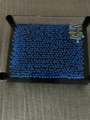 Blå pin art leksak i plast - Säljer en blå pin art leksak där du kan skapa 3D-former genom att trycka mot piggarna. Ramen är i svart plast och piggarna är blå. Perfekt för att göra roliga mönster och figurer med händer eller andra objekt.