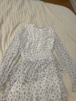 Vit blommig kortklänning Gina Tricot - Supersöt vit kortklänning från Gina Tricot med blått blommönster, v-ringning och långa ärmar. Klänningen har smock i midjan och volangdetaljer som ger en flowy känsla. Perfekt för sommarens alla tillfällen!