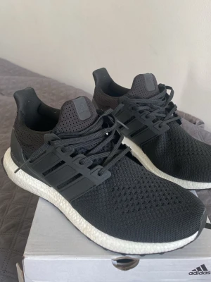 Svarta Adidas Ultraboost sneakers - Snygga svarta Adidas Ultraboost sneakers med stickad ovandel i mesh för bra andningsförmåga. Skorna har en vit Boost-mellansula och svart yttersula med gröna detaljer undertill. Klassisk tre-randig design på sidorna och låga i modellen.