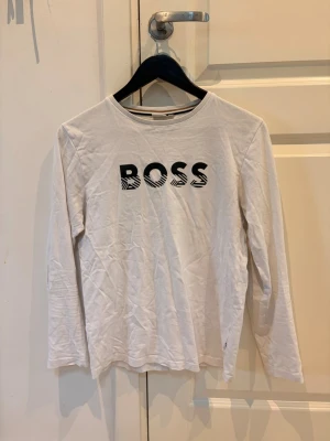 Vit långärmad topp från BOSS - Vit långärmad topp från BOSS med svart logotyp framtill i grafiskt mönster. Tillverkad i mjuk bomull med rund halsringning. Perfekt för en clean och stilren look.