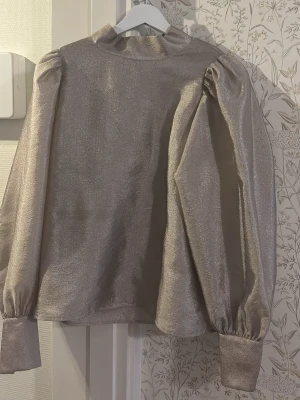 Champagnefärgad blus med puffärm - Elegant blus från Stockh LM i skimrande champagne/beige färg. Hög krage, puffärmar och dekorativ öppning med knappar i ryggen. Använd få gånger och i mycket fint skick. Passar perfekt till fest, middag eller mer uppklädda tillfällen.