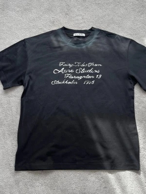 Svart t-shirt från Acne Studios - Svart t-shirt från Acne Studios med vit text på bröstet där det står 'Fairy Tales From Acne Studios Floragatan 13 Stockholm 1996'. Klassisk rund hals och normal passform. Perfekt för dig som gillar stilrena och trendiga plagg.