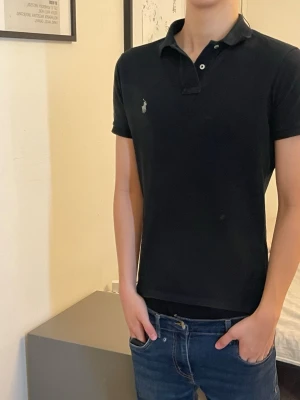 Ralph Lauren Svart pikétröja - Klassisk svart pikétröja från Ralph Lauren med korta ärmar och liten broderad logga på bröstet. Tröjan har krage och två knappar vid halsen. Perfekt till jeans eller chinos för en clean och stilren look. Som passar till alla typer av klädstilar