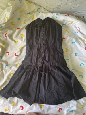 Svart kort klänning med snörning - Säljer en svart axelbandslös kort klänning med snygg snörning framtill. Klänningen har en lätt utställd kjol och är perfekt för dig som vill ha en edgy och trendig look. Materialet känns mjukt och följsamt, troligen bomull eller polyester. OBS!! De hårda ”pinnarna” (boning) i korsetten är böjda där fram men den är fortfarande fin på. Storlek M men går att snöra åt så den passar S