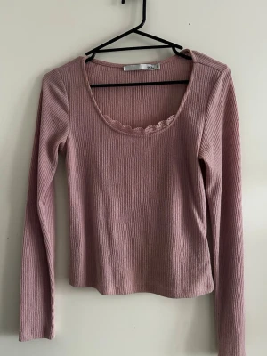 Ljusrosa ribbad långärmad topp - Söt ljusrosa ribbad topp med lång ärm och rundad urringning dekorerad med spetskant. Toppen har en croppad passform och är tillverkad i mjukt bomullsmaterial. Perfekt för dig som gillar en enkel men ändå gullig stil.
