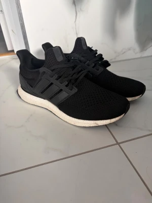 Adidas ultraboost 1.0 - Snygga svarta sneakers med vit, dämpad sula och ventilerande mesh-ovandel. Skorna har klassisk snörning och diskreta detaljer i svart plast längs sidorna. Perfekta för dig som gillar stilrena och bekväma sneakers till vardags.