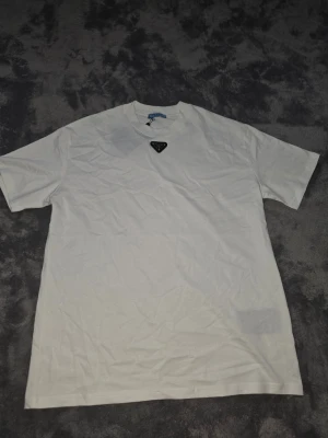 Vit t-shirt från Prada med metallogo - Säljer en clean vit t-shirt från Prada med en ikonisk metallogo på bröstet. Klassisk rund halsringning och korta ärmar. Perfekt basplagg med exklusiv känsla och stilren design.