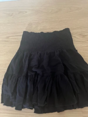 Svart volangkjol med smock - Snygg svart kjol med bred smock i midjan och flera volanger. Kjolen är kort och har en luftig känsla, perfekt för en trendig och avslappnad stil. Materialet ser ut att vara tunn och mjuk bomull eller viskos.