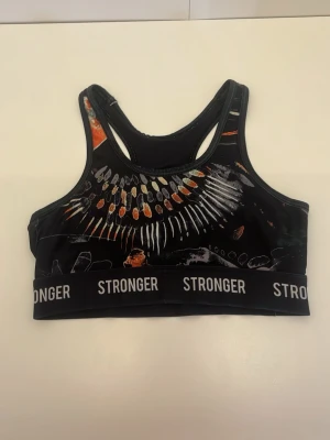 Mönstrad sport-bh från Stronger - Snygg sport-bh från Stronger med svart bas och färgglatt abstrakt mönster i orange, vitt och grått. Resårband med logga runt om och brottarrygg för extra stöd. Perfekt för träning och aktiva dagar.