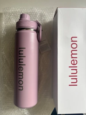 Ljus lila vattenflaska från Lululemon - Snygg ljus lila vattenflaska från Lululemon med praktiskt lock och handtag. Flaskan är tillverkad i rostfritt stål och har en stilren design med Lululemon-loggan tryckt längs sidan. Perfekt att ha med till gymmet, skolan eller på språng.
