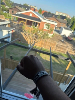 Grått Christian Dior tygarmband - Snyggt grått tygarmband från Christian Dior med vit text och dekorativa tofsar i ändarna. Armbandet har en justerbar knytning och ger en trendig och avslappnad look. Perfekt accessoar för att lyfta din outfit.