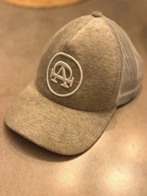 Grå meshkeps med broderad logga - Snygg grå keps med böjd skärm och vit broderad logga framtill. Baksidan är i meshmaterial för extra ventilation och avslappnad stil. Perfekt för dig som gillar streetwear och vill ha en enkel men stilren accessoar.