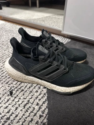 Svarta Adidas Ultraboost sneakers - Säljer ett par svarta Adidas Ultraboost sneakers med stickad ovandel och vita Boost-sulor. Skorna har klassiska tre ränder på sidan, svart snörning och en bekväm, sportig design. Perfekta för dig som gillar streetwear och sportig stil.