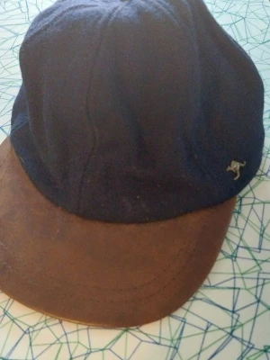 Mörkblå keps med brun skärm - Snygg keps i mörkblått tyg med en brun skärm i mockaimitation. Klassisk form med en liten metallkänguru på sidan som detalj. Perfekt för dig som vill ha en stilren accessoar med lite retrovibbar.