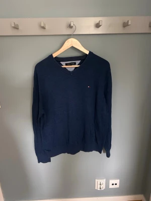 Mörkblå V-ringad tröja Tommy Hilfiger - Mörkblå tröja med V-ringning från Tommy Hilfiger. Tröjan har en liten broderad logga på bröstet och ribbade muddar vid ärmslut och nederkant. Tillverkad i mjukt material som ger en clean och stilren look.