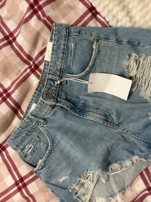 Ljusblå denimshorts med slitningar - Säljer ett par ljusblå denimshorts med råa kanter och slitna detaljer framtill och baktill. Shortsen har klassisk femficksdesign, knapp och dragkedja i gylfen samt medelhög midja. Aldrig använda 