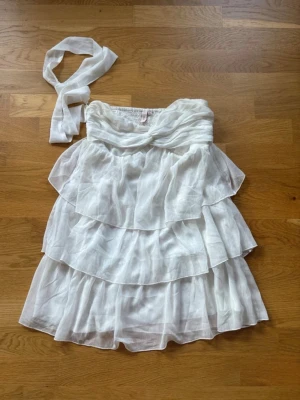 Vit omlottklänning från Nelly - Säljer en vit axelbandslös omlottklänning från Nelly i luftig och lätt polyester. Klänningen har flera lager volanger och medföljer ett matchande band som kan knytas i midjan eller runt halsen. Perfekt för sommarens alla tillfällen!