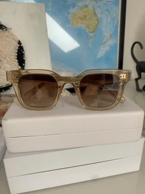 Beige fyrkantiga solglasögon CHIMI - Snygga solglasögon från CHIMI med transparent beige båge och fyrkantig form. Glasen är bruna och ger en cool retrovibe. Perfekta för soliga dagar och en stilren look.