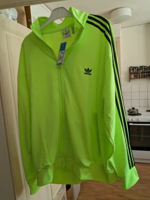 Neongrön Adidas ziptröja - Neongrön ziptröja från Adidas med svarta ränder längs ärmarna och Adidas-logga på bröstet. Tröjan har hög krage, dragkedja framtill och breda muddar vid ärmslut och nederkant. Perfekt för en sportig och retro vibe. Storlek XXXL
