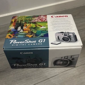 Canon PowerShot G1 digitalkamera - Canon PowerShot G1, en kompakt digitalkamera i silverfärg med svart objektivlock. Kameran har en klassisk, kantig design och levereras med originalkartong, laddare, rem och tillbehör. Perfekt för dig som gillar retrostil och vill ha en smidig kamera.
