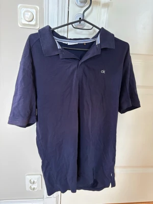 Mörkblå pikétröja Calvin Klein - Mörkblå pikétröja från Calvin Klein med klassisk krage och korta ärmar. Tröjan har diskret 'ck'-logga på bröstet och randiga detaljer på insidan av kragen i vitt och blått. Tillverkad i mjuk bomull, perfekt för en avslappnad stil.