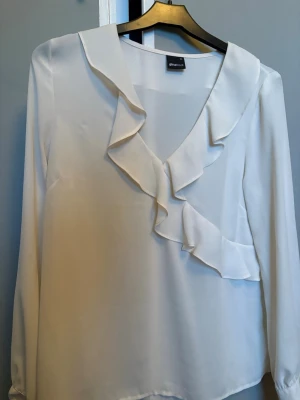 Vit blus med volang från Gina Tricot - Stilren vit blus från Gina Tricot med långa ärmar och snygga volangdetaljer längs den djupa V-ringningen. Nästa helt ny, använd 2 gånger😍