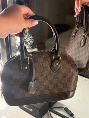 Brun handväska med rutigt mönster - Snygg handväska i brunt med klassiskt rutigt mönster och mörkbruna detaljer. Väskan har två handtag, guldfärgade metalldetaljer och dragkedja upptill. Materialet ser ut att vara konstläder eller canvas. Perfekt storlek för det viktigaste och ger en lyxig känsla.
