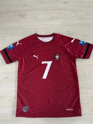 Portugal matchtröja Ronaldo #7 - Säljer en röd Portugal matchtröja från Puma med vita detaljer och vågmönster. Tröjan har nummer 7 och Ronaldo på ryggen, samt landslagets emblem på bröstet. Tillverkad i funktionsmaterial (Drycell) och har korta ärmar.