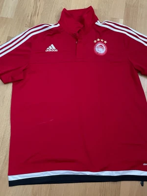 Röd Adidas Olympiakos träningströja - Röd träningströja från Adidas med Olympiakos-emblem och fyra stjärnor på bröstet. Tröjan har vit logga, vita ränder på axlarna och vit kant nertill. Tillverkad i funktionsmaterial (climacool) och har kort dragkedja vid halsen.