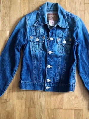 True religion jeansjacka modell Jimmy storlek S - True religion jeansjacka med långa ärmar, markerad krage och silverfärgade knappar framtill. Två bröstfickor med lock och knappdetaljer. Storlek small. Modell Jimmy. Jackan har en cool vintagekänsla och tydliga sömmar som ger extra karaktär. Perfekt att slänga över en hoodie eller t-shirt.  Mått: Axel till ärmslut: 62 cm Axel till nederkant: 54 cm Midjan/nederkant: 40 cm  Mycket fint skick.