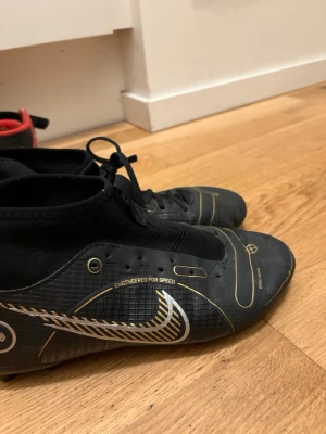 Svarta Nike fotbollsskor med guld - Säljer ett par svarta Nike fotbollsskor med gulddetaljer och vita inslag. Skorna har högre strumpkrage för extra stöd runt ankeln och snörning för bra passform. Sulan har dobbar för spel på gräs. Snygg design med Nike-logga på sidan.