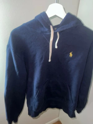Mörkblå hoodie från Polo Ralph Lauren - Mörkblå hoodie från Polo Ralph Lauren med klassisk gul logga broderad på bröstet. Hoodien har huva med vita dragsnören och en stor magficka. Tillverkad i mjukt bomullsmaterial och har ribbade muddar vid ärmar och nederkant.