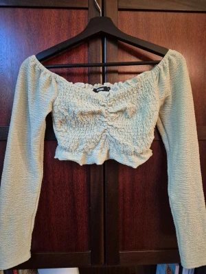 Grön croppad offshoulder topp - En pastellgrön croppad topp med offshoulder och långa ärmar. Toppen har smockat tyg och en rynkning framtill som ger en snygg look. Perfekt att styla med jeans eller kjol en somrig look.