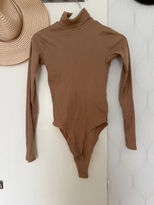 Beige ribbad body med polokrage - Snygg beige body med lång ärm och polokrage. Tillverkad i ett mjukt, ribbat material som sitter tajt mot kroppen. Perfekt att styla med jeans eller kjol för en clean look. Enkel att knäppa i grenen och har en neutral färg som passar till mycket.