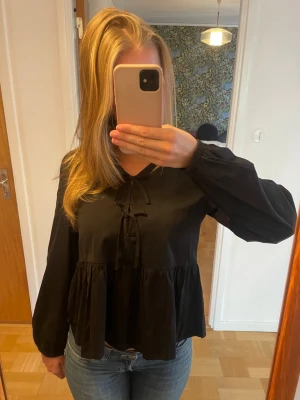 Svart volangblus - Snygg svart blus med volang nedtill och knytband vid halsen. Blusen har långa och lite puffiga ärmar. Aldrig använd. 