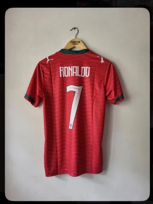 Röd Portugal fotbollströja Ronaldo - Säljer en röd Portugal fotbollströja med Ronaldo och nummer 7 på ryggen. Tröjan har gröna detaljer vid krage och ärmslut samt vita tryck. Perfekt för dig som älskar fotboll och vill stötta ditt favoritlag.