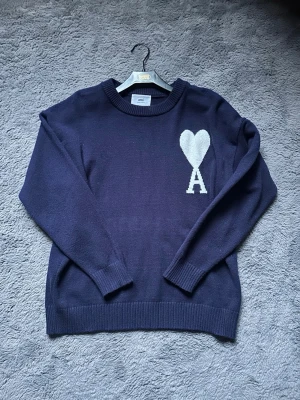 Ami Paris stickad tröja - Mysig mörkblå stickad tröja med vit hjärtdetalj och bokstaven A på bröstet. Tröjan har rund halsringning, ribbade muddar och lång ärm. Perfekt för dig som gillar enkel men snygg design.   Använd ett fåtal gånger, inga defekter. Passar både small och M